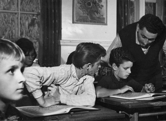 "Schule" - Schüler haben vor sich einen Atlas aufgeschlagen, ein Lehrer beugt sich über das Buch eines Jungen; um 1970