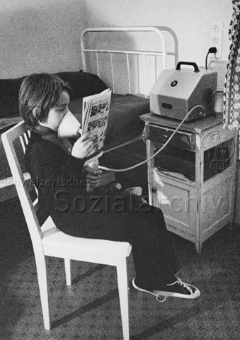 "Kinderheilstätte [Davos], Therapie - Junge sitzt auf einem Stuhl, hält sich ein Atemgerät vor Mund und Nase und liest einen Comic; 1971