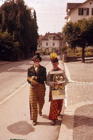 "Kinderzirkus/ Kindertheater: Kinderzirkus Robinson" - Zwei als Clowns verkleidete Jungen, einer mit Tschinellen,d er andere mit Flugblättern und einem Plakat des "Wanderzirkus Buchegg"; um 1970