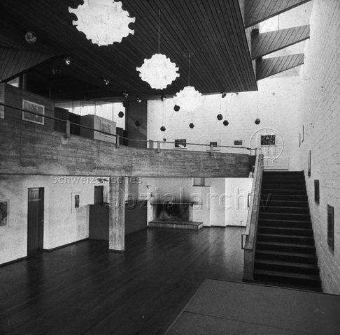 "Freizeitanlagen, Gemeinschaftszentren: Saal FZA Heuried, Zürich" - Blick in einen grossen Raum mit Holzboden und Cheminée, eine Treppe führt in den ersten Stock, in dem die Räume von einer Galerie aus zugänglich sind; um 1970