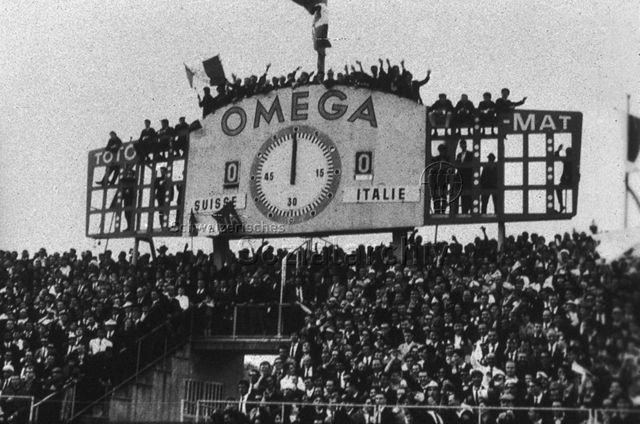 Fussballländerspiel Schweiz - Italien: Blick auf eine überfüllte Tribüne mit einer Grossanzeige von einer Uhr, die auf 12 Uhr steht und Spielstand  Null zu Null, zahlreiche Männer in Anzügen sind auf die Anzeigetafel geklettert, winkend und jubelnd; um 1965