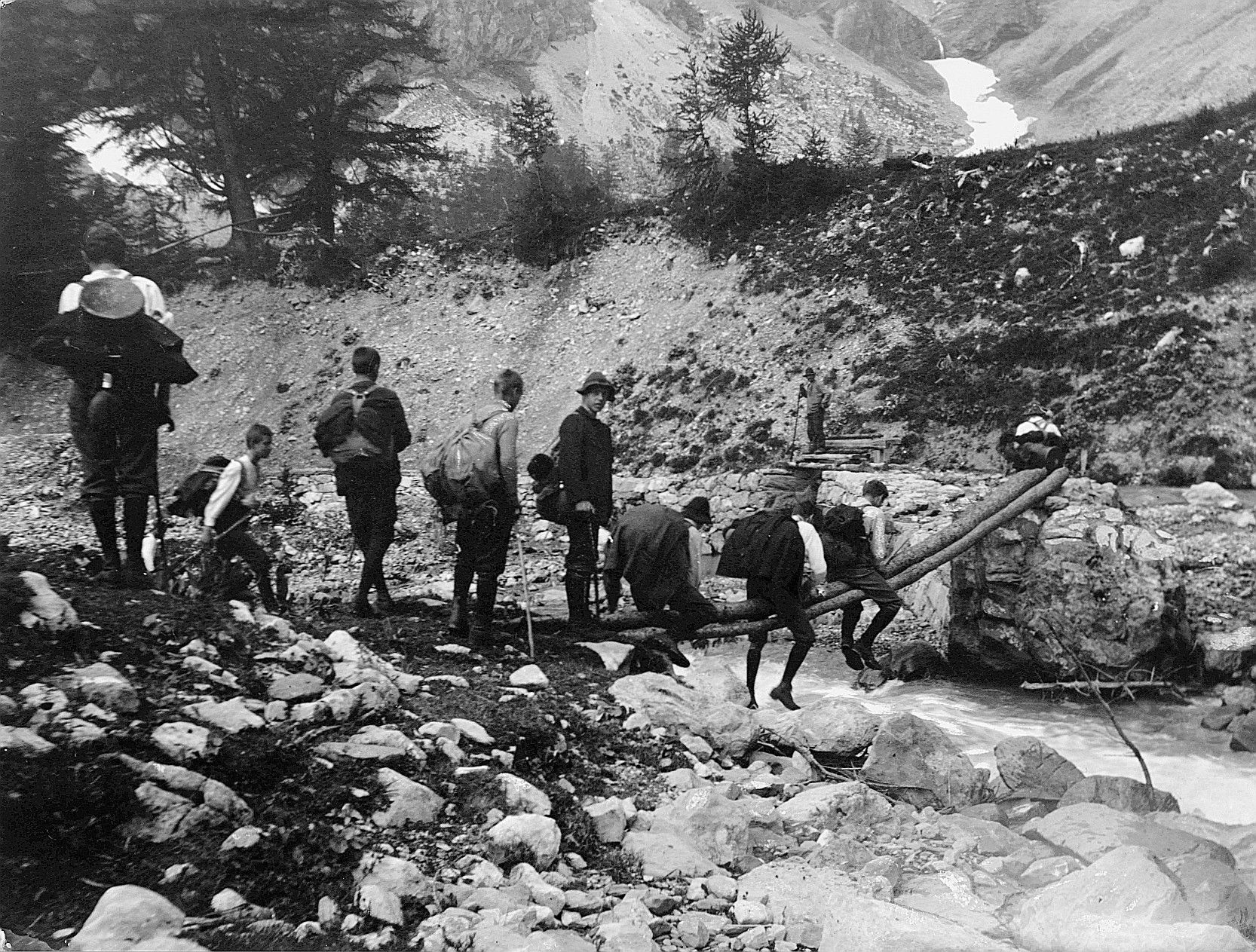 Wandervögelswanderung in den Bergen - Drei Wandervögel sitzen rittlings auf zwei Baumstämme, welche als Brücke für die Überquerung e