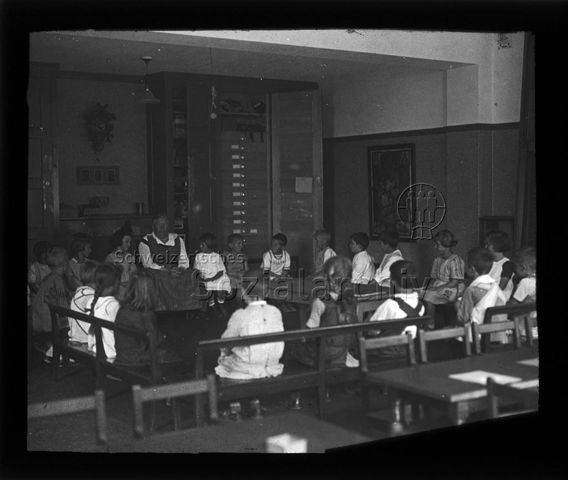 Diaserie zum Thema Kindergarten / Schulkind; Kinder und eine Frau sitzen in einem Schulzimmer auf Holzbänken im Kreis; um 1930