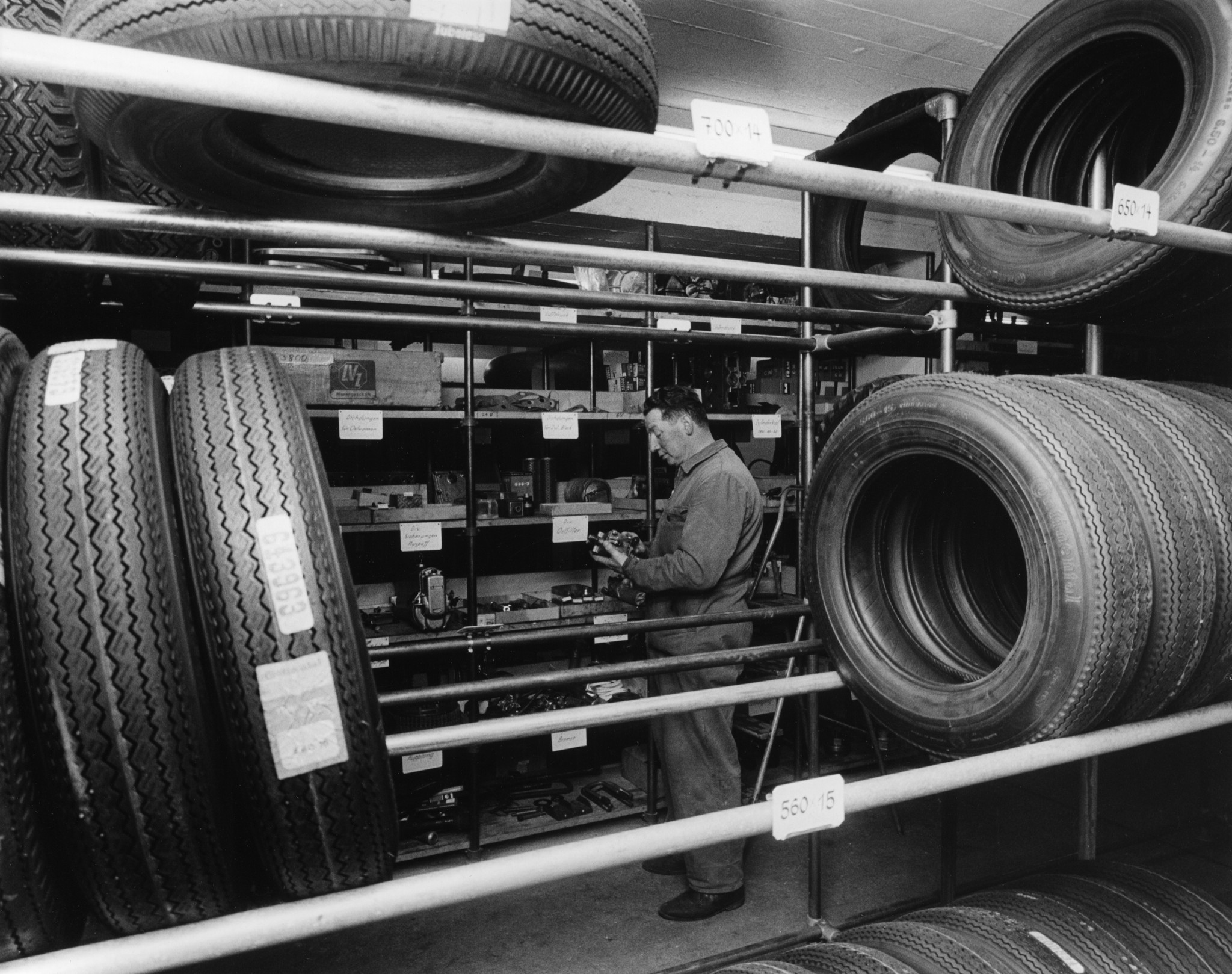 Automechaniker beim Begutachten eines Ersatzteils vor einem Gestell, im Vordergrund gelagerte Autoreifen