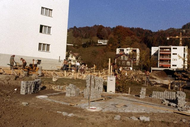 "Siedlungs- und Quartierspielplätze: Fronarbeit Siedlungsspielplatz, Ebnat-Kappel SG" - Baustelle des Kinderspielplatzes auf einer grossen Wiese, es entsteht ein Platz aus Pflastersteinen; um 1970