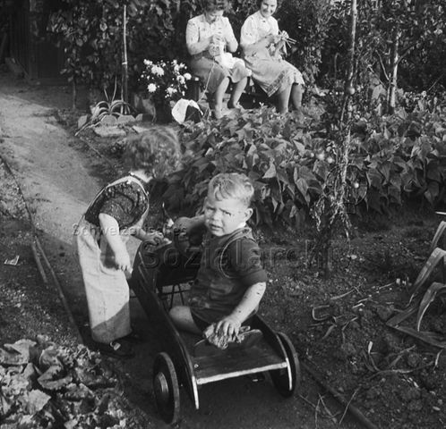 "Kleine Kinder spielen im Garten" - Junge und Mädchen mit einem Tretauto, hinten sitzen zwei Frauen auf einer Bank und stricken; um 1960