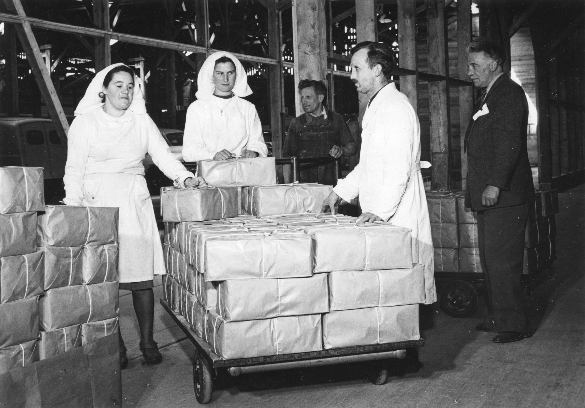 "Verpackungslager des Colis Suisse in Genf"; zwei Schwestern und ein Arzt (?) sowie zwei weitere Männer stehen um ein Palett mit Paketen