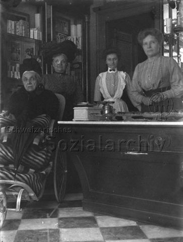 Vier Frauen (u.a. Frau im Rollstuhl, Verkäuferin) in einem Geschäft mit Süssgebäck und einer Vitrine mit Schokolade, um 1920