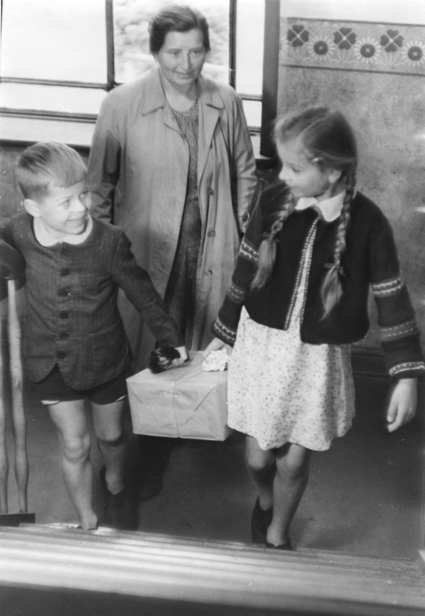 "Hamburg, Sommer 1948: ein Freipaket kommt an!"; zwei Kinder tragen vor ihrer Mutter ein Paket die Treppe hoch