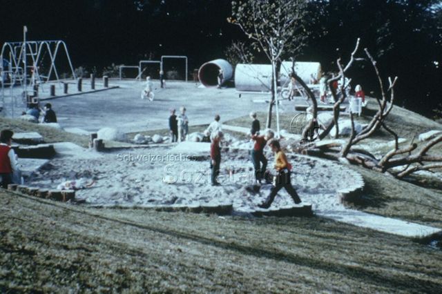 "Siedlungs- und Quartierspielplätze: Siedlungsspielplatz Adliswil ZH" - Blick auf den Spielplatz, Kinder spielen im Sandkasten/ bei den Steinen/ auf dem Hartplatz; um 1970