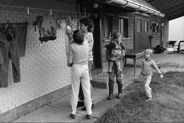 "Praktikantenhilfe" - Eine Praktikantin und vier Kinder beim Wäscheaufhängen; um 1980