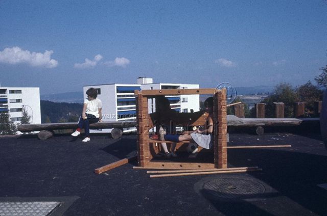 "Siedlungs- und Quartierspielplätze: Siedlungsspielplatz Adliswil ZH" - Zwei Kinder sitzen in einem aus zusammensteckbaren Holzteilen gebauten Häuslein auf dem Hartplatz; um 1970