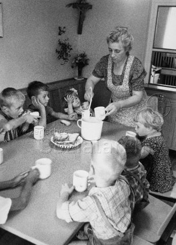 "Heimkinder, SOS-Kinderdorf" - Betreuerin schöpft Suppe, Kinder sitzen am Tisch; um 1965