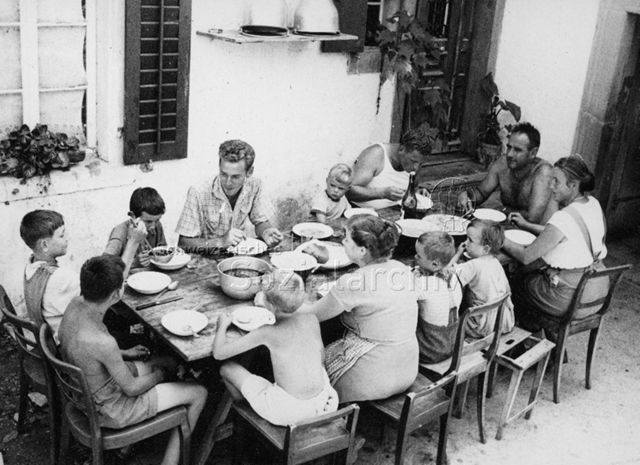 PraktikantInnenhilfe - Praktikant und zwei Familien sitzen am Tisch und essen; um 1965