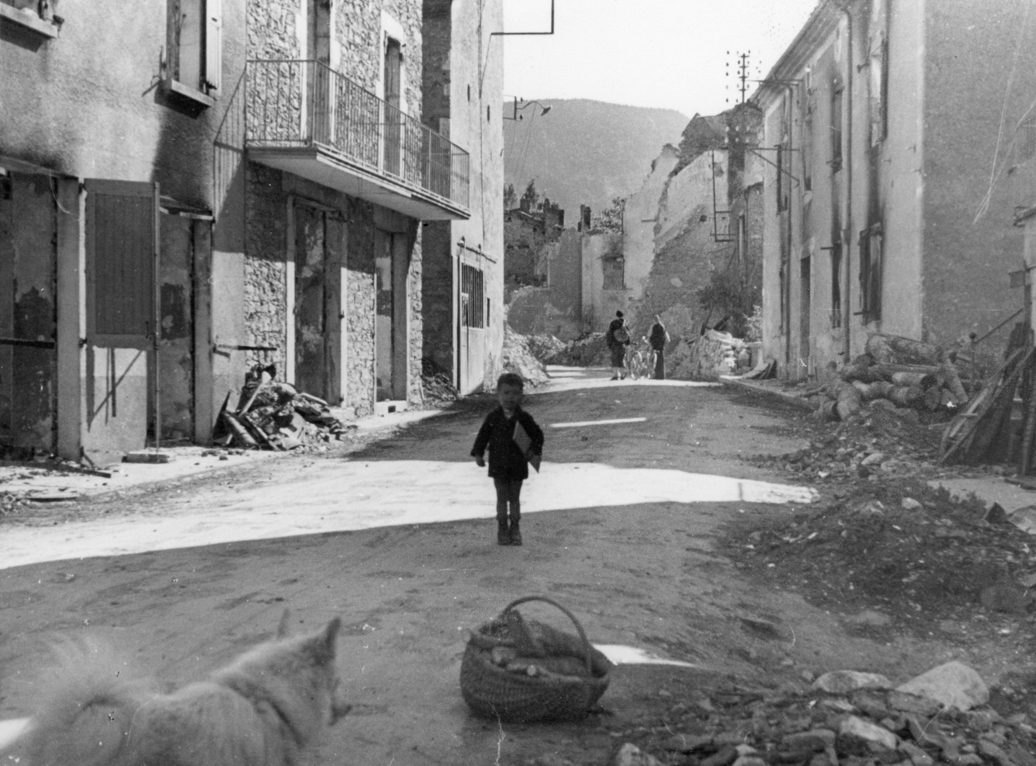 "Parmi les ruines"; ein Knabe steht auf einer Strasse, die von den Trümmern der Häuser gesäumt ist