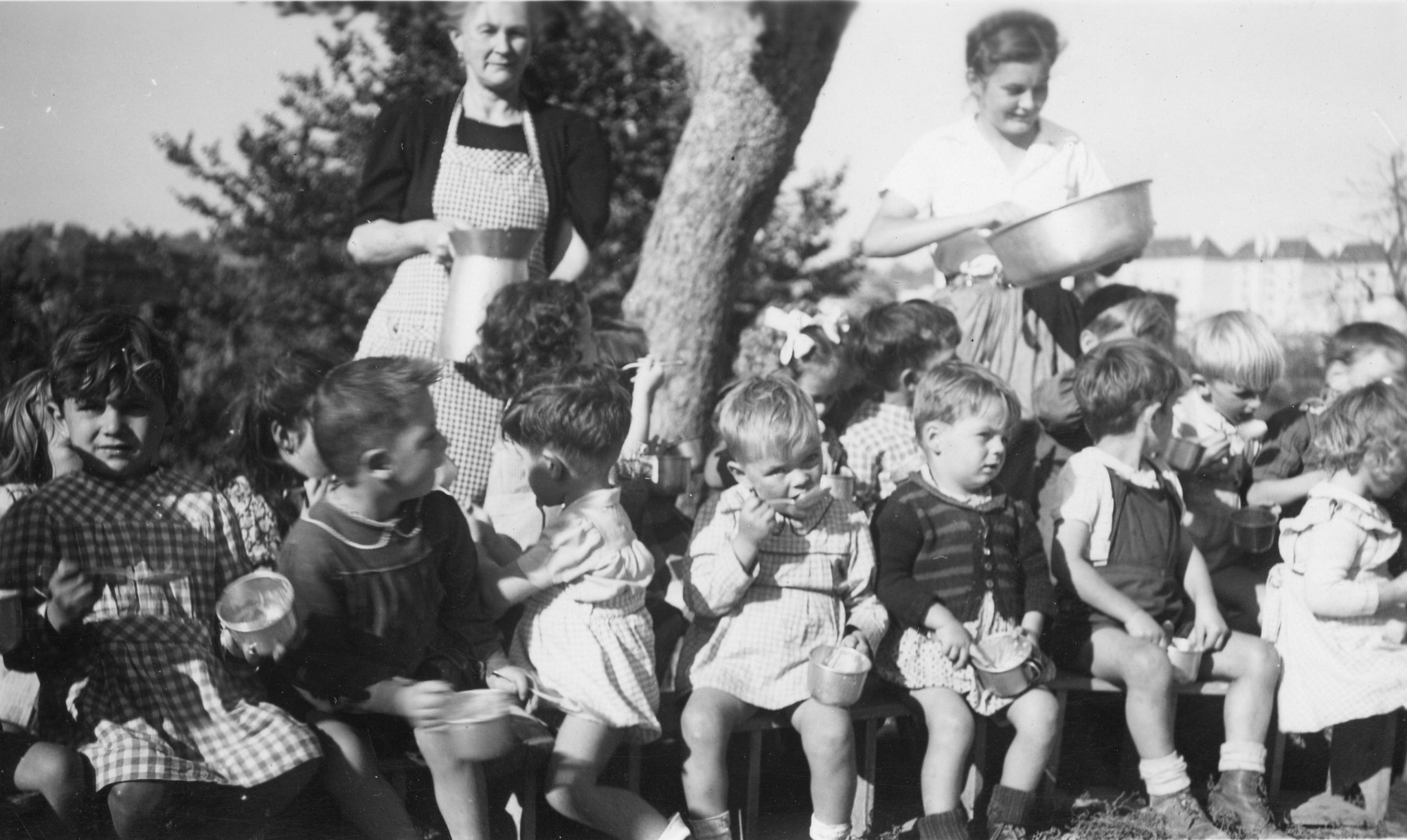 Kinder sitzen in einem Kreis um einen Baum und essen aus Schüsselchen; im Hintergrund verteilen zwei Betreuerinnen das Essen
