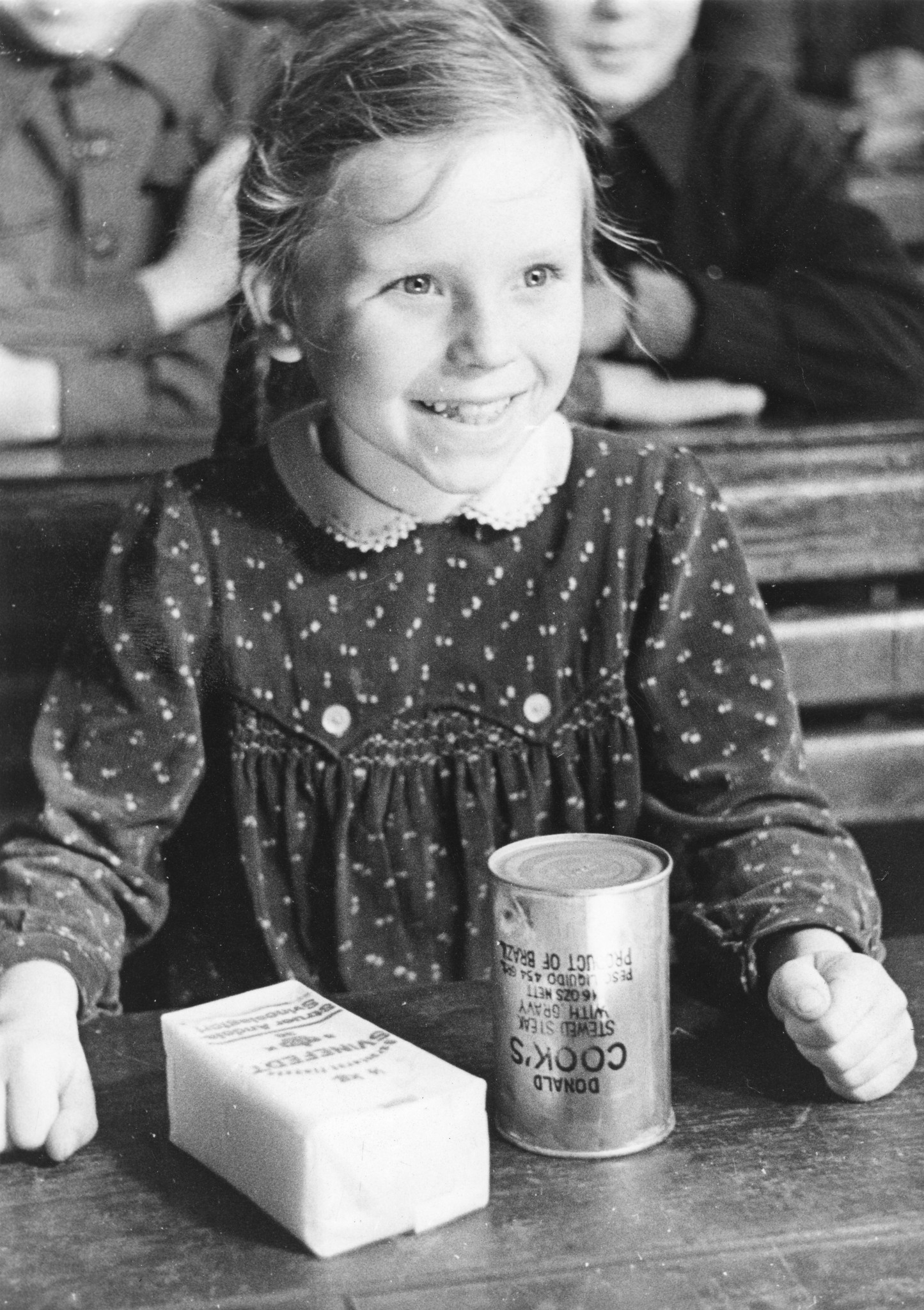 "März 1948: Verteilung von Lebensmittelpaketen am die Schüler der Gemeinschaftsschule Düsseldorf"; ein Mädchen lachend vor einer Dose Fleischeintopf und einer Packung Schweinefett sitzend