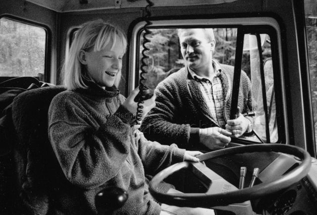 Kinder besuchen ihre Eltern am Arbeitsplatz - Tocher am Funkgerät eines Fahrzeugs, Vater steht draussen; um 1995