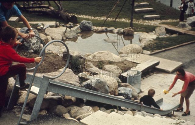"Siedlungs- und Quartierspielplätze: Rutschbahn Siedlungsspielplatz Adliswil ZH, 1970" - Kinder am Rutschen, eine Mutter fängt ihr Kind auf, im Hintergrund sieht man einen kleinen Teich