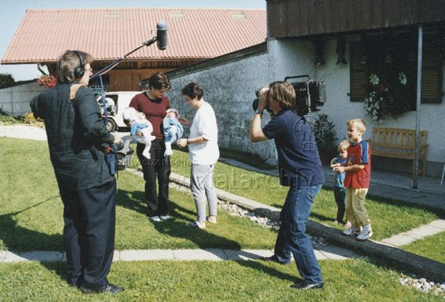 PraktikantInnenhilfe: Einsätze - Mutter und Praktikantin mit zwei Säuglingen werden von Fernsehteam gefilmt, daneben stehen zwei Kinder; 2000