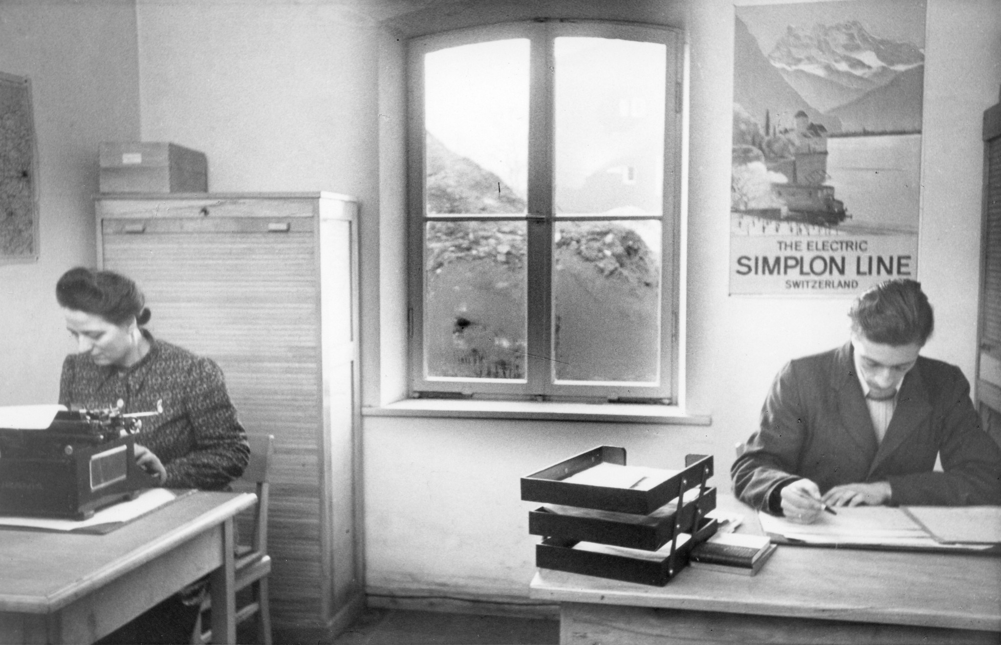 Zwei Büroangestellte sitzen an ihren Plätzen und arbeiten; im Hintergrund hängt ein Plakat mit der Aufschrift "The electric Simplon Line Switzerland"