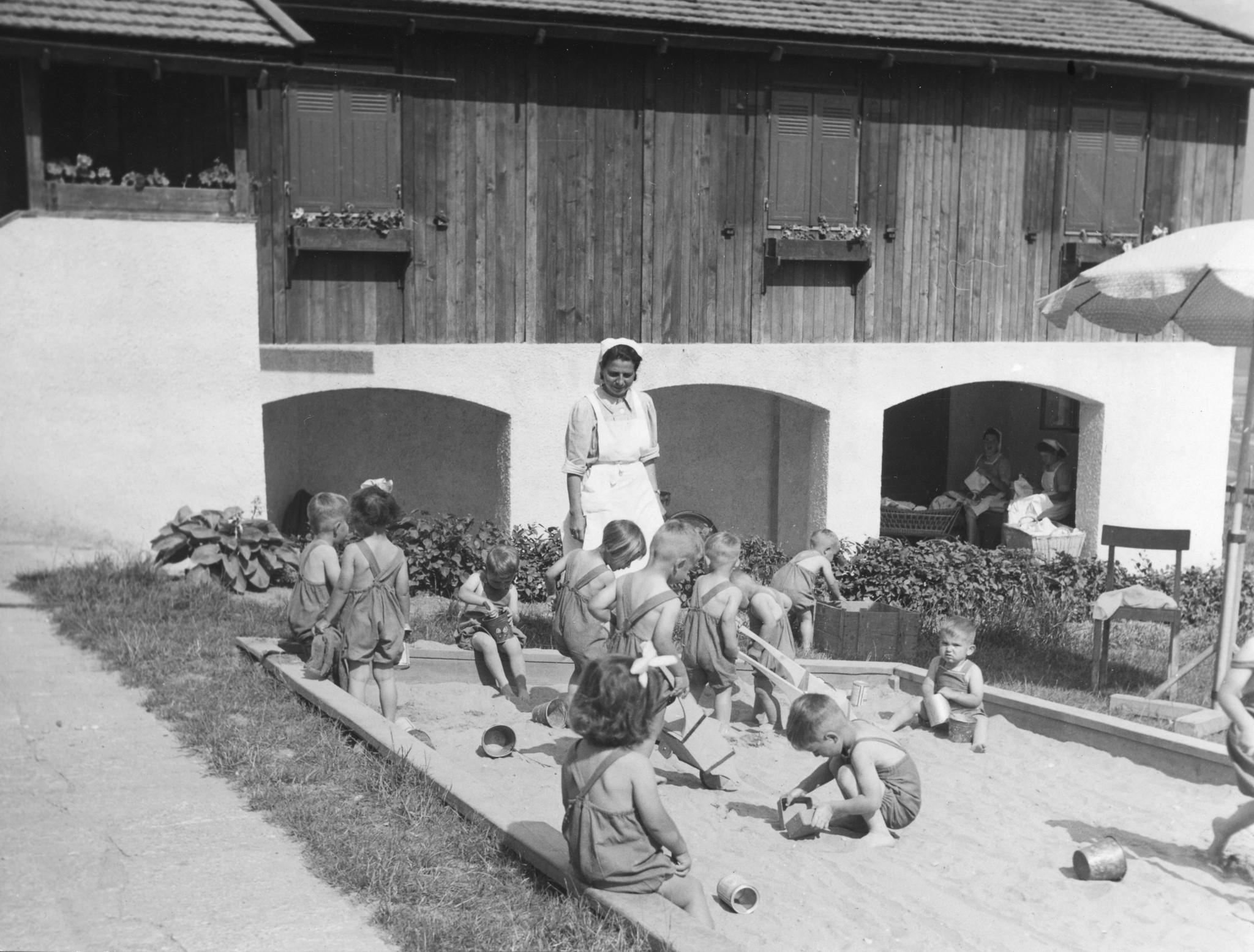 "Schwyzerhüsli Juni. 50"; Kleinkinder spielen im Sandkasten vor einem Haus