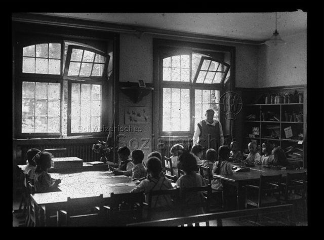 Diaserie zum Thema Kindergarten; Kinder sitzen in einem Schulzimmer an zwei Tischen; Kinder am ersten Tisch weben mit Papier, Kinder am zweiten Tisch arbeiten mit Holzklötzen; Betreuerin am Kopfende des zweiten Tisches stehend; um 1930
