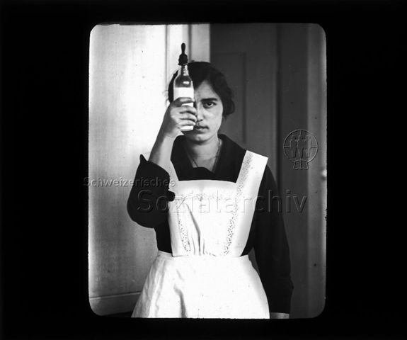 Diaserie zum Thema Säuglingspflege; Krankenschwester eine Säuglingsflasche vor ihr rechtes Auge haltend; um 1930